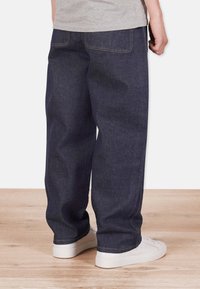 Sininen farkkuhousu, joissa on rento istuvuus, takataskut, näkyvät ompeleet ja suora lahje, yhdistettynä valkoisiin lenkkareihin.