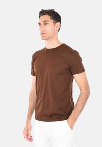 T-shirt marrone a maniche corte in tessuto morbido, con scollatura a girocollo e forma aderente, indossato con pantaloni bianchi.