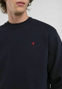 Felpa girocollo blu navy realizzata in morbido tessuto, con un colletto a costine e un logo ricamato rosso sul lato sinistro del petto.