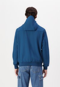 Blå zip-jacka med hög krage, som har elastiska mudd och midja. Konstruerad av ett glatt tyg, baksidan visar rena linjer och minimala accenter.