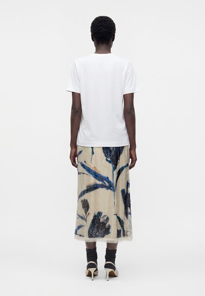 Personne portant un t-shirt blanc uni à manches courtes, une jupe beige avec un motif floral bleu, des chaussettes noires et des chaussures à talons hauts beiges, debout de dos.