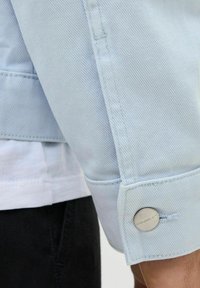 Svijetloplava traper jakna s vidljivim šavovima, detaljem gumba i blago teksturiranim materijalom. Crna košulja ispod.