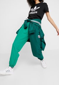 Black T-shirt with white Adidas logo, paired with green track pants featuring side stripes and ankle cuffs. White sneakers with colored accents.