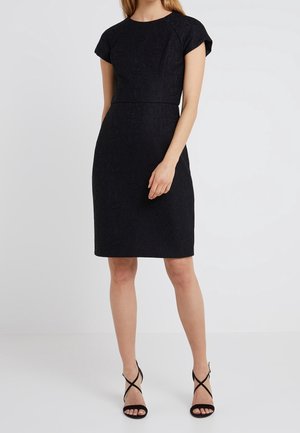 Kobieta w dopasowanej czarnej sukience do kolan z krótkimi rękawami oraz w czarnych sandałkach na wysokim obcasie z paskami, stojąca na jednolitym tle.