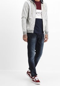 Man som bär en grå zip-up hoodie, en färgblockerad t-shirt i marinblått och vinrött, mörka jeans, röda och vita sneakers, med tatuerade händer, stående på vit golv.