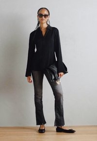 Schwarzes Langarmhemd mit Knopfverschluss, graue Schlagjeans, schwarze Ballerinas und eine schwarze Handtasche, accessorisiert mit übergroßen Sonnenbrillen.