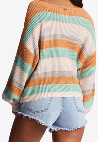 Strickpullover mit horizontalen Streifen in Orange, Blaugrün und Beige, mit weitem Ausschnitt und lockerer Passform, kombiniert mit ausgefransten Jeansshorts.