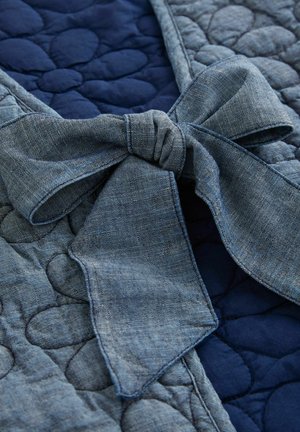 Denim mašna na tamnoplavom, teksturiranom, kviltanom materijalu s okruglim oblicima, koja prikazuje kombinaciju materijala i boja.