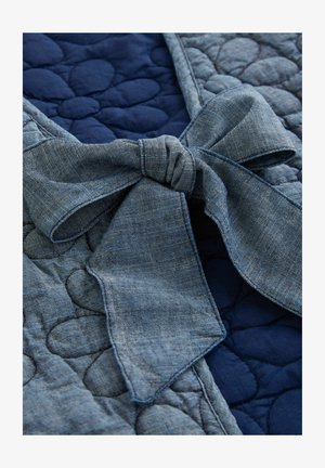 Denim-Schleife über einem strukturierten, dunkelblauen, gepolsterten Stoff mit runden Formen, der eine Mischung aus Materialien und Farben zeigt.