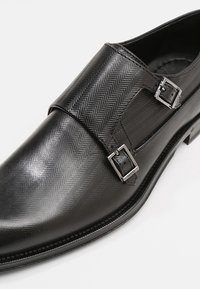 Chaussure habillée en cuir noir avec un motif en chevrons, deux boucles, et un design de bout légèrement pointu avec une finition lisse.