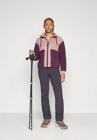 Naine kannab kahte tooni lilla jope ja tumedaid pükse, hoides kahte musta matkapulka, seistes ühtlase heledal taustal.