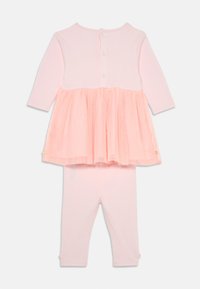 Roza dvodelna obleka, ki vključuje srajco z dolgimi rokavi in tulle krilo preko ter ustrezne legice. Gumbi na hrbtni strani in sijoči detajli.