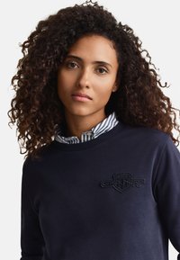 Göndör hajú nő, aki tengerészkék pulóvert visel csíkos ing gallérral alatta, közvetlenül a kamerába néz.