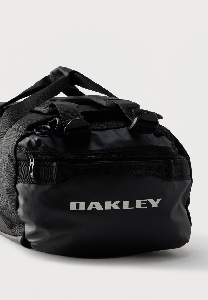 Sort Oakley sportstaske med lynlåslomme og dobbelt bærehåndtag på hvid baggrund.