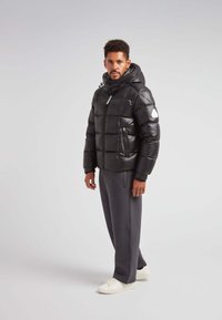 Giacca a vento nera con finitura lucida, modello trapuntato orizzontale, con zip frontale e cappuccio. Accentuata con un logo bianco sul braccio.