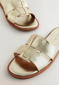 Flade sandaler med skinnende metallisk guld materiale, brede stropper med vævet design og en brun gummisål.