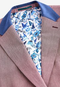 Hellbrauner Blazer mit blauem Kragen, der ein floral gemustertes Innenfutter in Blau und Weiß aufweist. Texturiert, modernes Design.