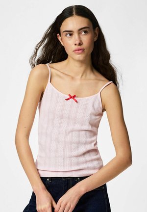 Jeune femme aux longs cheveux foncés portant un haut rose clair sans manches avec un nœud rouge et un jean foncé, posant sur un fond blanc.