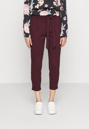 Vrouw draagt korte bordeauxrode broek met een gestrikte tailleband, bloes met lange mouwen en witte instapschoenen, staand op een lichte vloer.