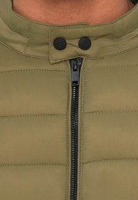 Chaqueta de plumas en verde oliva con un cuello alto, dos cierres de botón negro y una cremallera frontal con herrajes negros y costuras visibles.