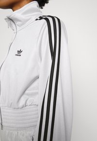 White athletic jacket with a high collar, elasticized waistband, and black three-stripe detailing along the shoulders. Features Adidas logo.