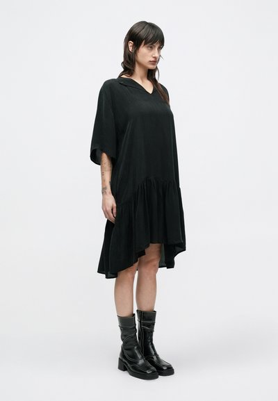Μαύρο φόρεμα με V-λαιμόκοψη, κοντά μανίκια και βολάν στο τελείωμα. Το ύφασμα φαίνεται λείο και ρέον. Συνδυάζεται με μαύρες μπότες μέχρι το γόνατο.