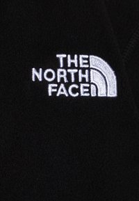 Černá fleecová tkanina s bílým vyšívaným logem "THE NORTH FACE" s zakřiveným designovým prvkem, na pozadí jemné textury.