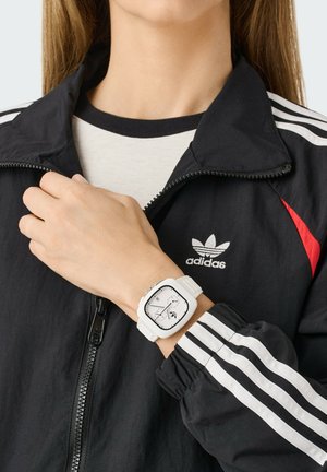 Orologio sportivo bianco con quadrante quadrato, indicatori delle ore neri e cinturino in silicone bianco, indossato con una giacca nera con zip caratterizzata da dettagli e strisce rosse.