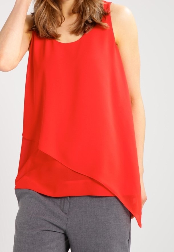 Donna con top asimmetrico rosso senza maniche e pantaloni grigi, con capelli castani sciolti che le cadono sulle spalle.