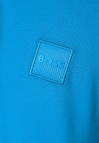 Tissu texturé bleu vif avec un patch carré cousu présentant un texte en relief "BOSS" en fil bleu assorti.