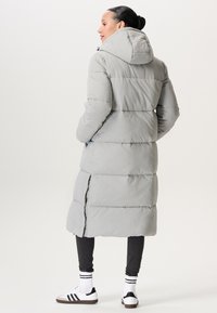 Vrouw in een lange lichtgrijze pufferjas met een capuchon, donkere leggings, witte sokken met zwarte strepen en witte sneakers, die van opzij staat.