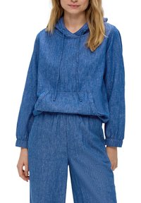 Vrouw in een blauwe denim-look hoodie met trekkoorden en bijpassende wijde broek, staand met een ontspannen houding.