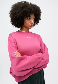 Rozā oversize džemperis ar apaļu kakla izdobi un garām, apjomīgām piedurknēm. Uz krūtīm ir neliels balts Nike logo.