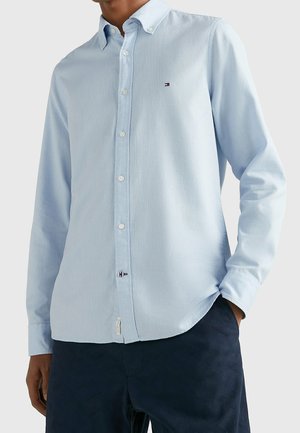 Mężczyzna ubrany w jasnoniebieską koszulę na guziki z długim rękawem, z małym logo na kieszeni na piersi, w ciemnoniebieskich spodniach, z ręką w kieszeni.