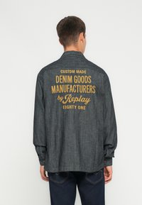 Camicia grigia in denim a maniche lunghe con vestibilità rilassata, caratterizzata da un ricamo giallo sul retro che recita "FABBRICANTI DI PRODOTTI IN DENIM SU MISURA di Replay."