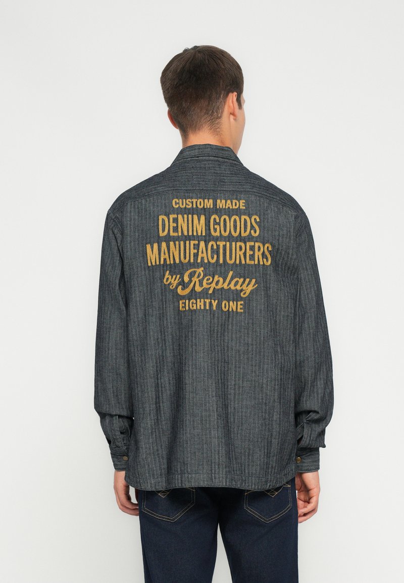 Camicia grigia in denim a maniche lunghe con vestibilità rilassata, caratterizzata da un ricamo giallo sul retro che recita "FABBRICANTI DI PRODOTTI IN DENIM SU MISURA di Replay."