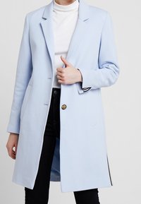 Cappotto lungo blu chiaro indossato sopra un dolcevita bianco e pantaloni neri, con una mano che sistema il cappotto vicino al petto.