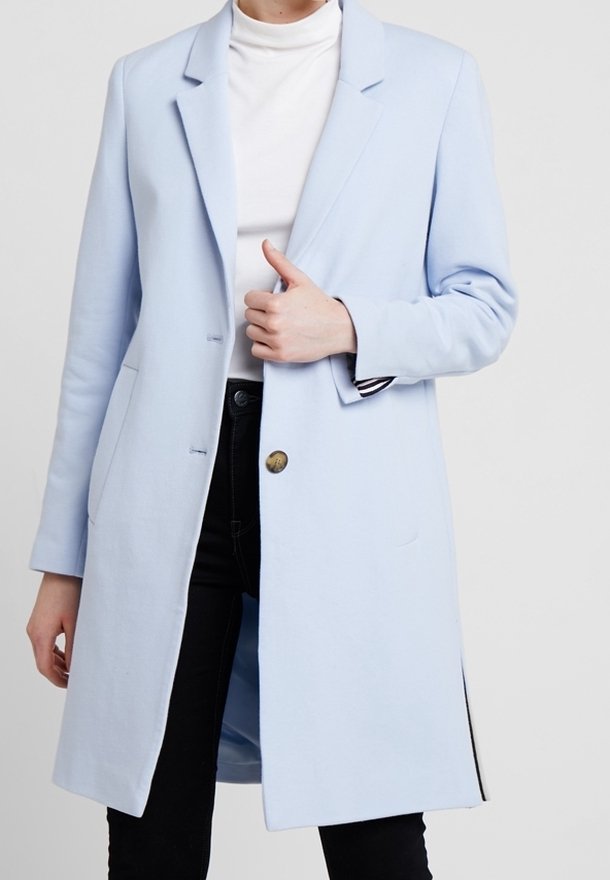 Cappotto lungo blu chiaro indossato sopra un dolcevita bianco e pantaloni neri, con una mano che sistema il cappotto vicino al petto.
