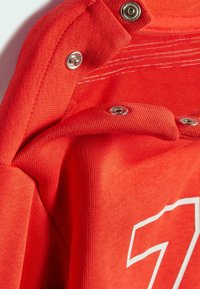 Primo piano di un tessuto rosso di un indumento con bottoni a pressione metallici e una stampa bianca con il numero 7. La texture è liscia con dettagli a motivo a maglia.