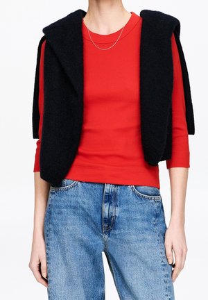 Personne portant une chemise rouge à manches longues avec un pull noir drapé sur les épaules et un jean bleu, visible du cou jusqu'aux hanches.