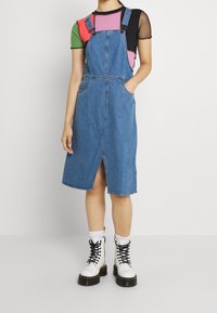 Denim hängselklänning med justerbara band, framfickor och en central slits. Bärs över en flerfärgad kortärmad topp och i kombination med vita stövlar.