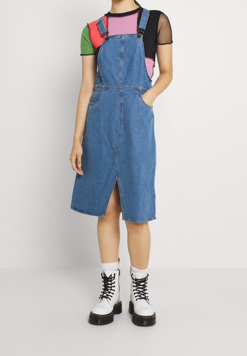 Denim hängselklänning med justerbara band, framfickor och en central slits. Bärs över en flerfärgad kortärmad topp och i kombination med vita stövlar.