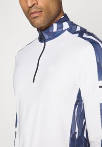 Haut à manches longues blanc avec un col haut, présentant des accents à motifs bleu marine sur les épaules et les côtés, fermeture éclair au niveau du cou.