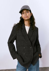 Blazer de cuadros gris oscuro de doble botonadura con solapas con muesca y bolsillos frontales, llevado sobre una blusa clara y combinado con jeans azules.