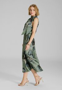 Abito verde in satin con stampa floreale, design senza maniche, dettaglio con fiocco al collo, gonna fluente, abbinato a sandali dorati con strappi.