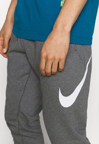 Pantalones de chándal grises hechos de material suave, con un logo blanco de Nike en el lateral y bolsillos laterales. La camisa es de color teal y tiene un diseño casual.