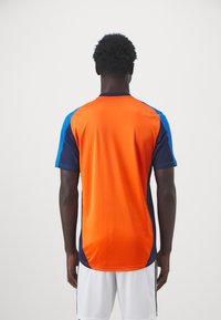 Camisa atlética com corpo laranja, mangas azuis e detalhes em azul-marinho. Feita de um tecido suave, apresentando mangas curtas e um design de gola redonda.