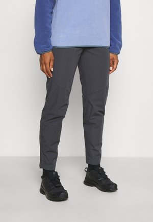 Persoon draagt donkergrijze broek, zwarte wandelschoenen, zwarte sokken en een blauw fleece top met lange mouwen en staat tegen een effen achtergrond.