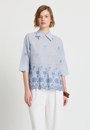 Frau trägt eine hellblaue Bluse mit Knöpfen und Blumenstickerei sowie weiße Hosen und steht vor einem schlichten Hintergrund.