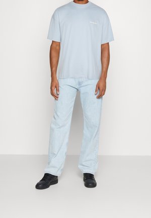 T-shirt en coton bleu clair avec col rond et manches courtes, associé à un jean denim clair et des chaussures noires. Fond neutre.